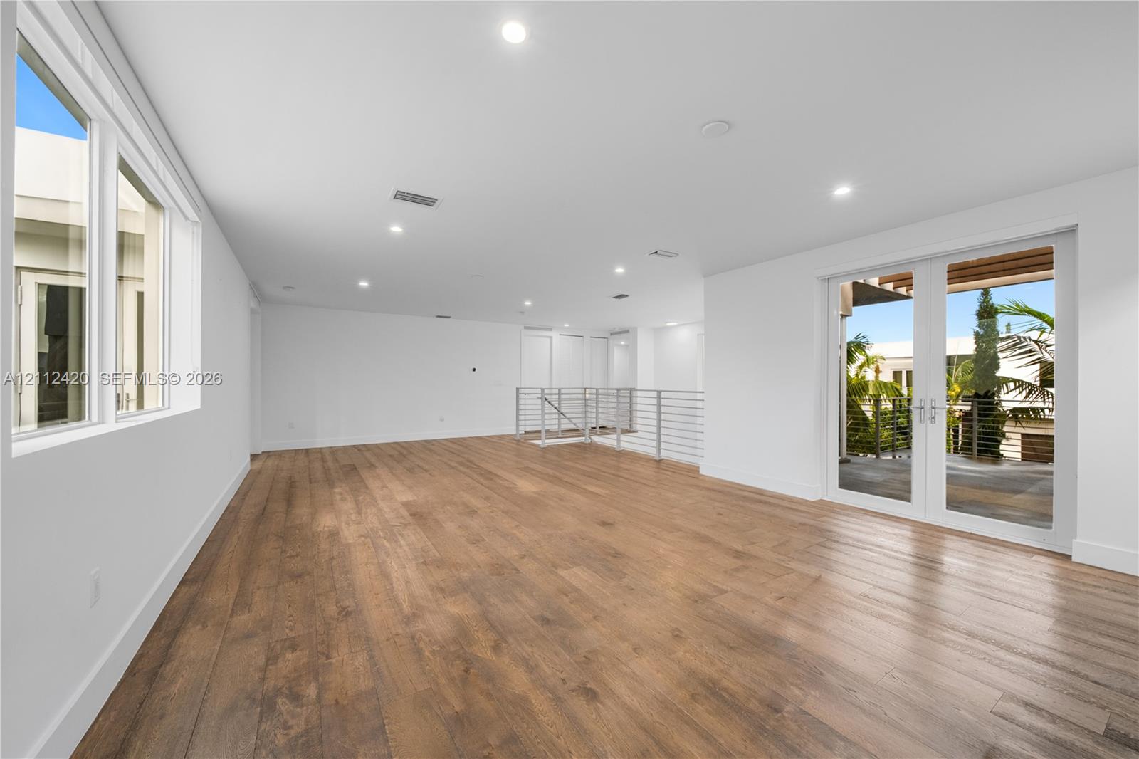 Open floor plan interior featuring wood-finish flooring, recessed lighting, multi-pane windows, and a metal horizontal-bar railing, with glass-paned double doors providing outdoor access