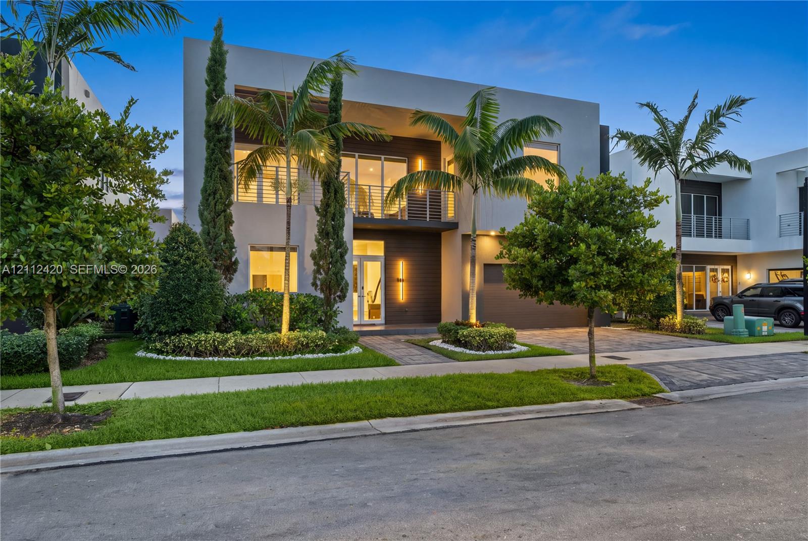 Modern two-story exterior featuring a smooth stucco faade, horizontal wood-style cladding, expansive glass panels, a second-level balcony with metal railing, and paver driveway