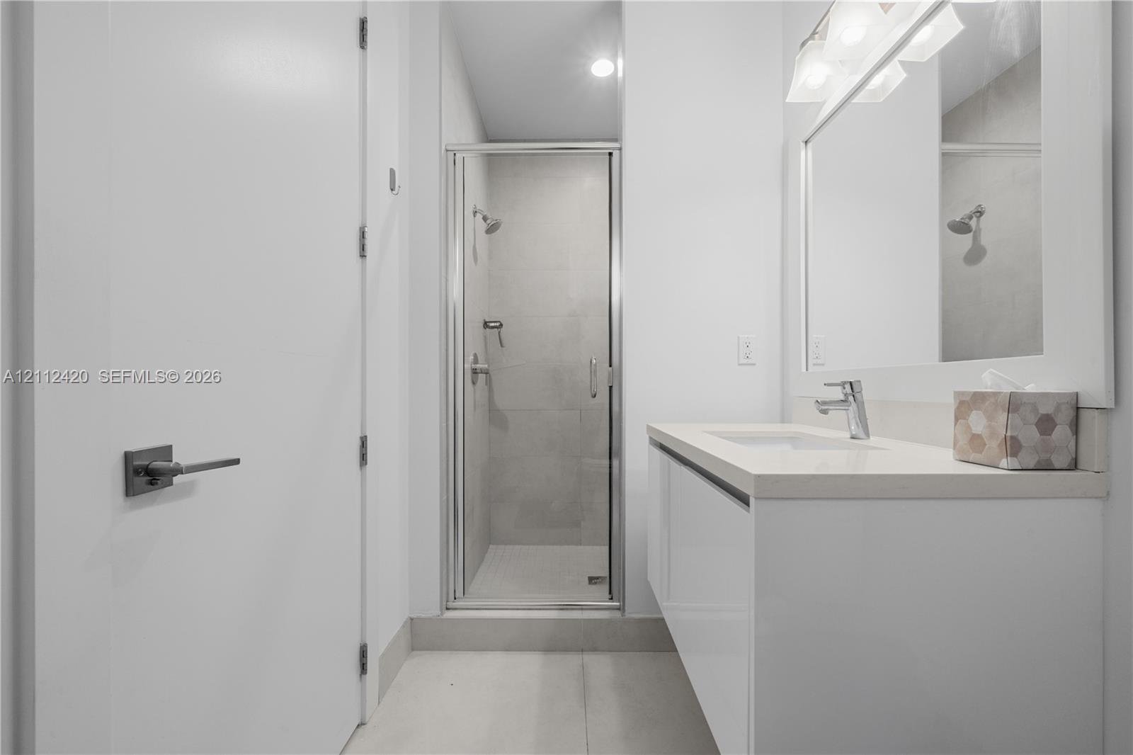 Modern vanity featuring a stone countertop and integrated sink, chrome faucet, framed mirror with mounted lighting, and a glass-enclosed shower with tiled walls
