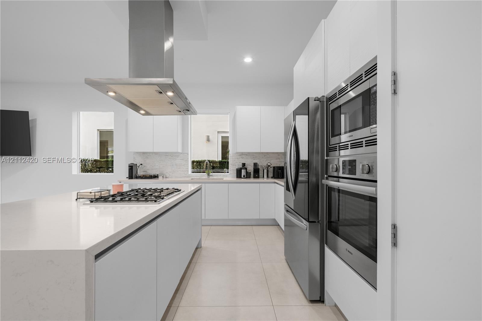 Contemporary kitchen featuring handleless white cabinetry, solid-surface countertops, a stainless steel range hood, built-in stainless steel double ovens, and tile flooring