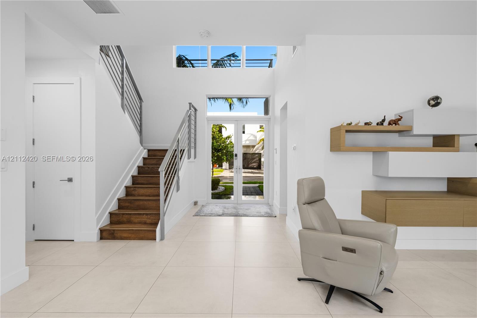 Entryway featuring a double-height ceiling, glass-paneled double front doors, transom windows, a staircase with wood treads and horizontal metal railings, and built-in wall-mounted cabinetry Entryway featuring a double-height ceiling, glass-paneled double front doors, transom windows, a staircase with wood treads and horizontal metal railings, and built-in wall-mounted cabinetry