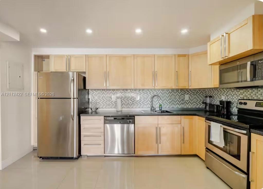 Kitchen featuring light wood cabinetry, stainless steel appliances, mosaic tile backsplash, dark countertops, and recessed lighting