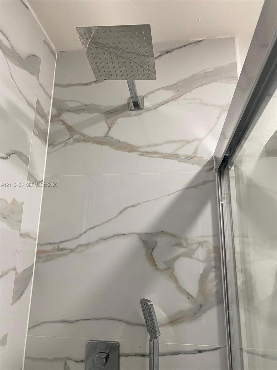 Walk-in shower featuring large-format wall tile with veining, a rainfall showerhead, a handheld shower wand, and a glass door enclosure