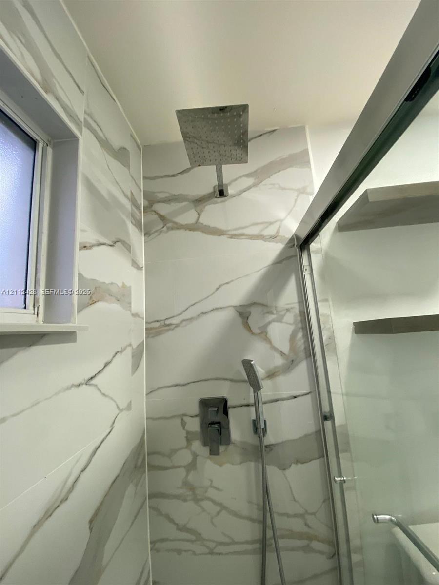 Walk-in shower featuring large-format wall tile with veining, a rainfall showerhead, a separate handheld sprayer, and built-in wall shelving enclosed by a glass door
