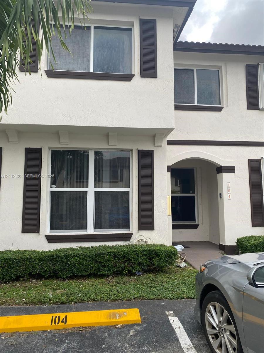 Two-story exterior featuring a light-toned stucco facade, dark decorative window shutters, an arched entryway, and manicured foundation shrubbery