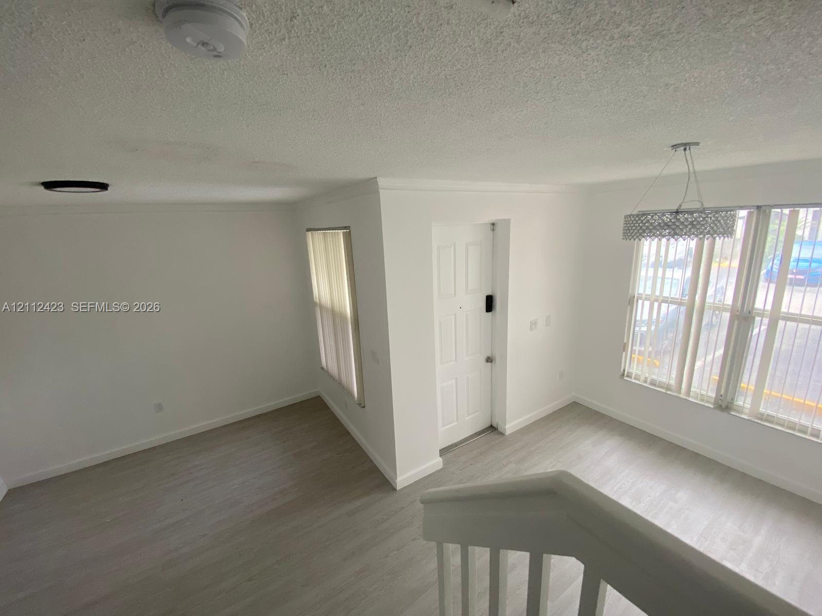 Neutral interior featuring wood-finish flooring, white walls, vertical window blinds, and a drum-style chandelier with beaded accents