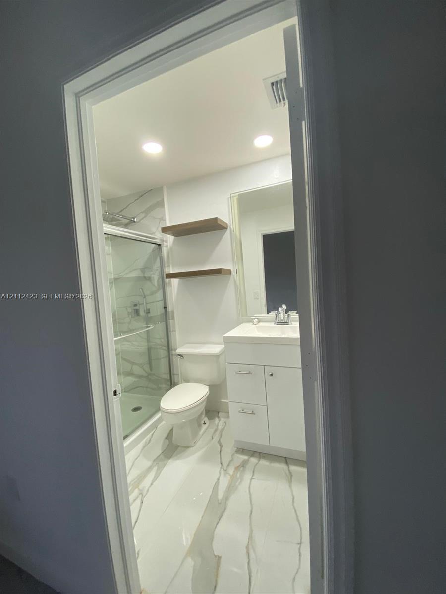Bathroom featuring a glass-enclosed shower with tiled surround, stone-finish flooring, white vanity with integrated basin, recessed lighting, and wall-mounted wood-finish shelving