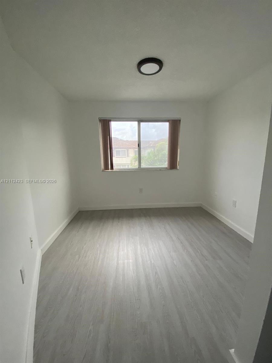 Neutral-toned interior space featuring wood-finish flooring, white walls, a centered window with vertical blinds, and a circular flush-mount ceiling light fixture