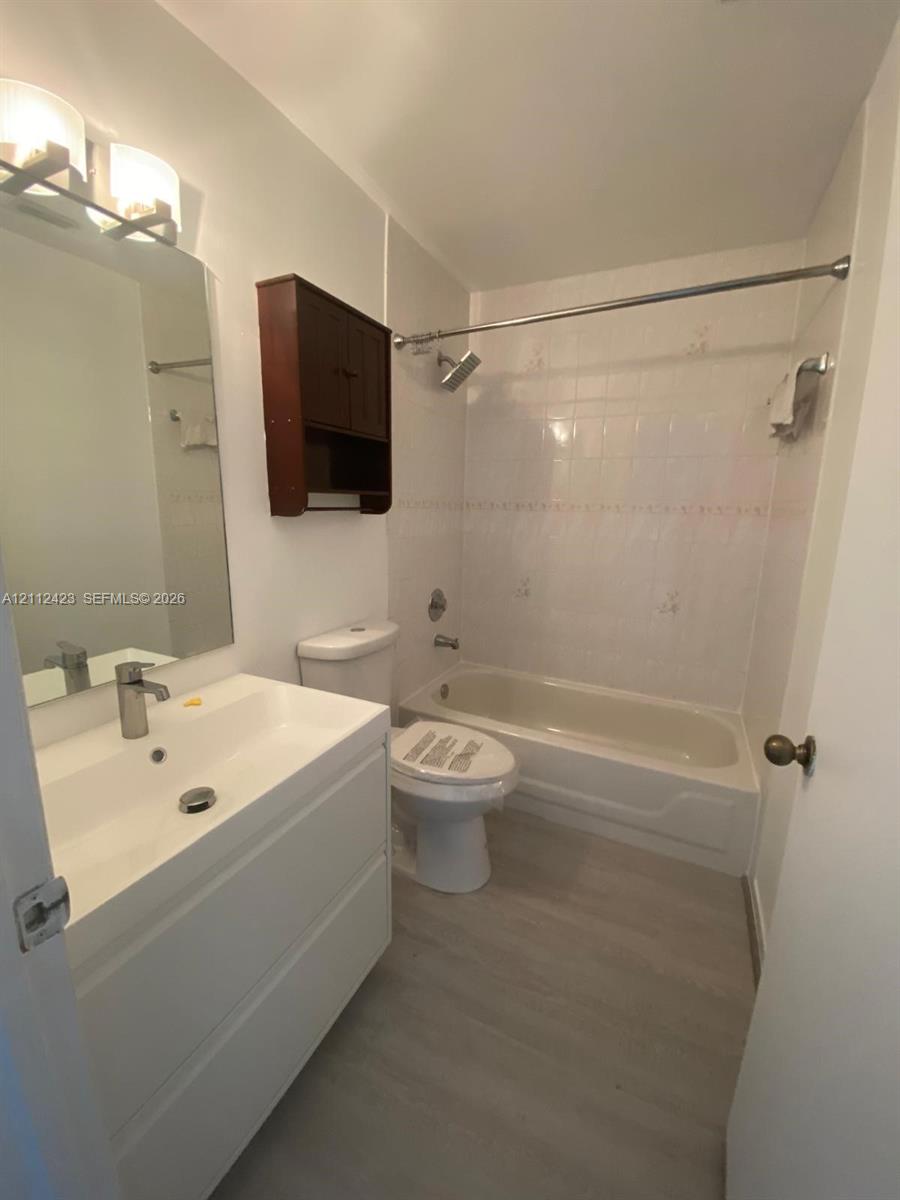 White vanity with integrated sink, rectangular wall-mounted mirror, light-toned wood-finish flooring, wall-mounted cabinet, and porcelain-tiled tub enclosure