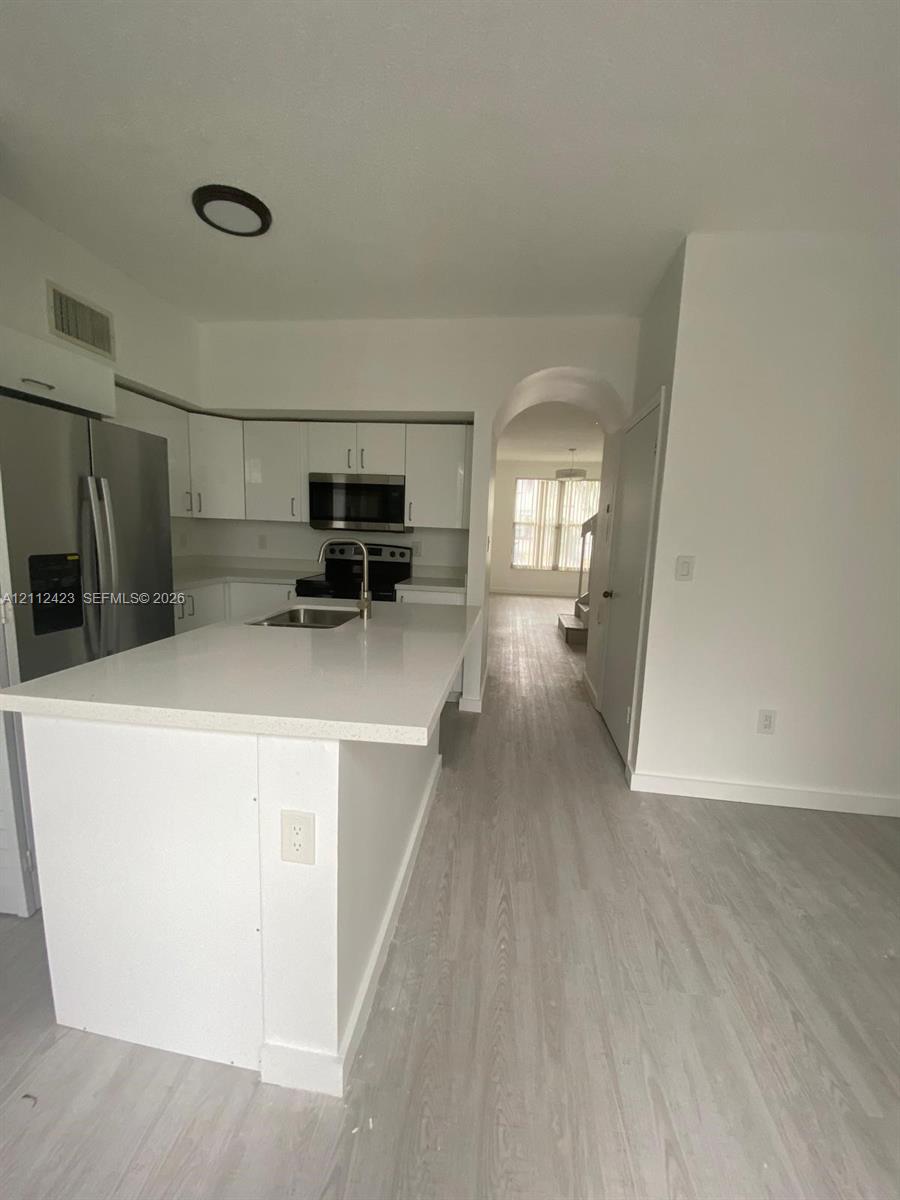 Kitchen featuring a stone-surface island, stainless steel refrigerator, flat-panel white cabinetry, wood-finish flooring, and an arched entry