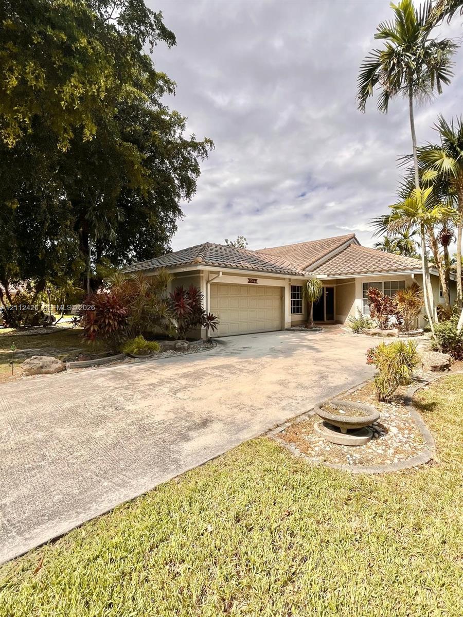 Single-story residence featuring a tile roof, an attached two-car garage, an expansive concrete driveway, and established palm and tropical landscaping Single-story residence featuring a tile roof, an attached two-car garage, an expansive concrete driveway, and established palm and tropical landscaping