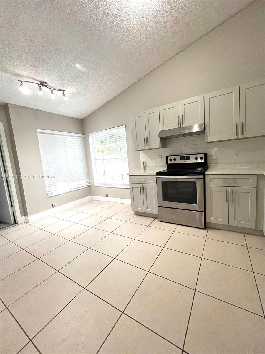 Kitchen featuring vaulted ceilings, light-tone cabinetry, stainless steel range with overhead vent hood, white tile backsplash, and oversized floor tiles