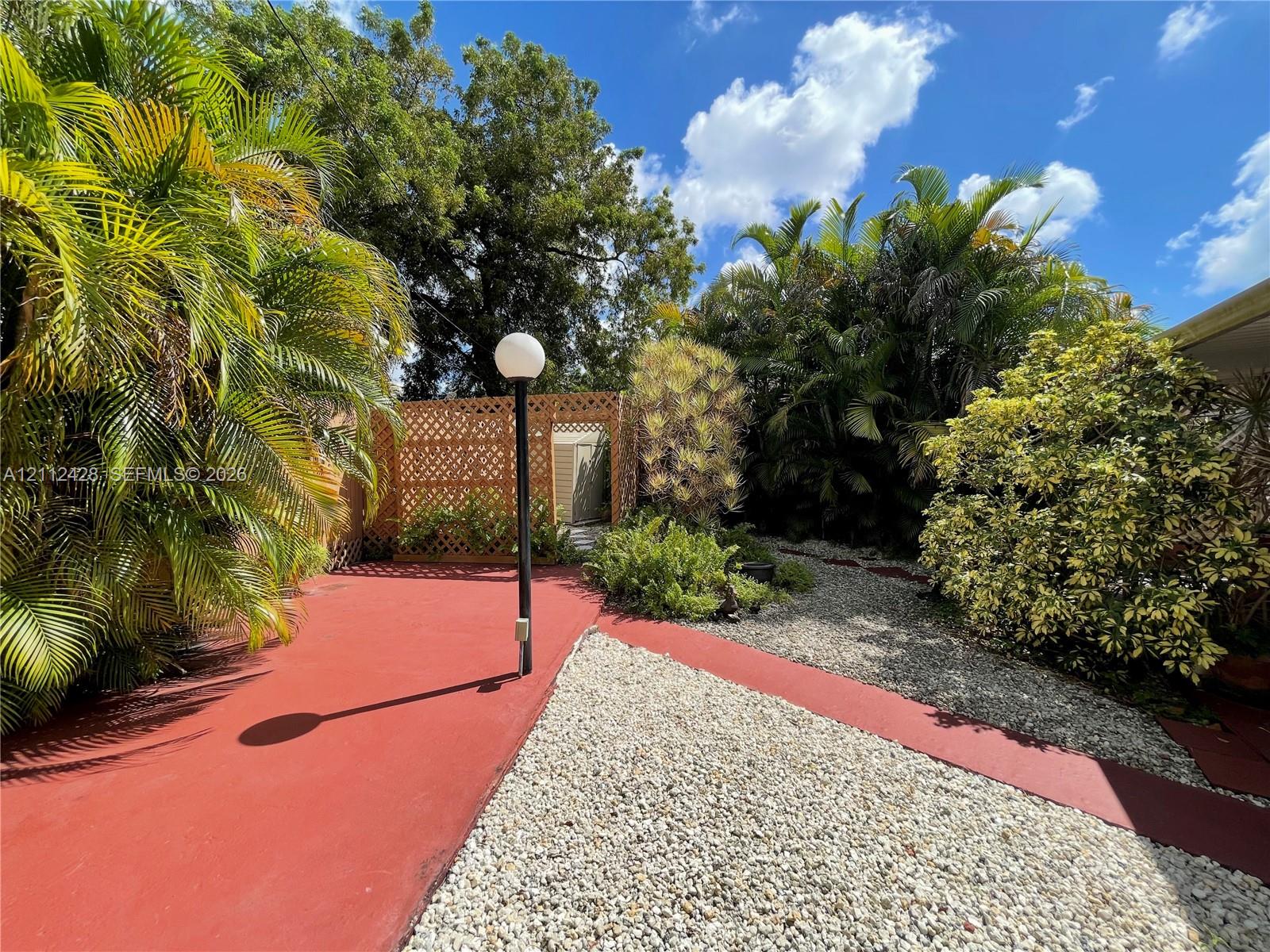 Outdoor space featuring a painted concrete patio, stone ground cover, lattice privacy screening, and mature tropical landscaping with an integrated lamp post