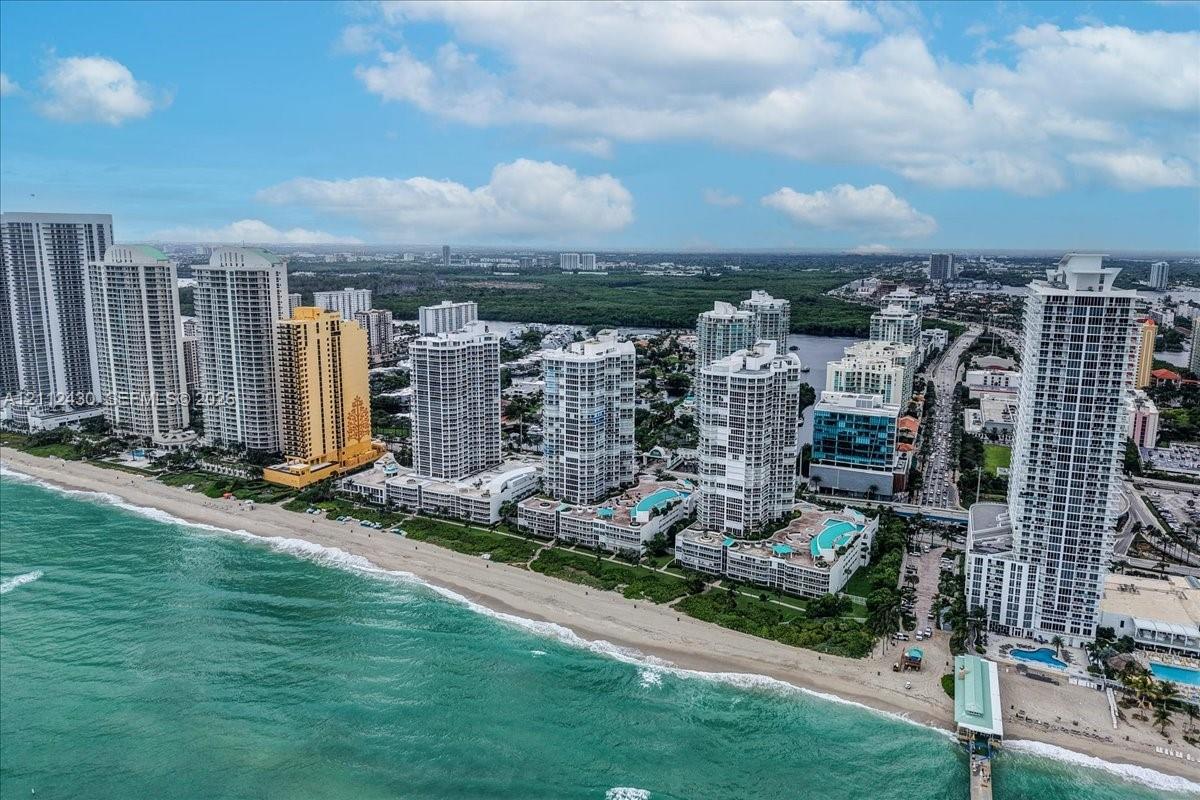 Aerial perspective of the coastline featuring oceanfront high-rise structures, sandy beach access, and tiered outdoor swimming pools