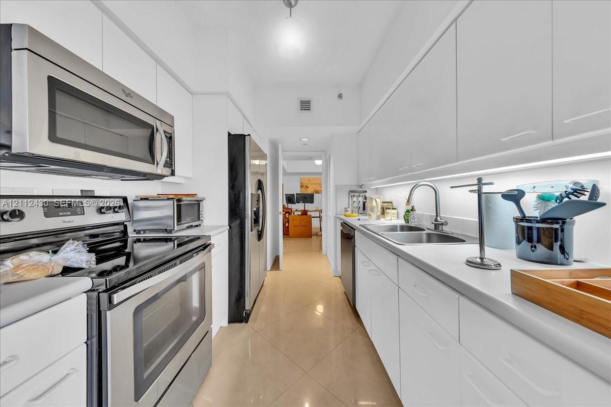 Galley kitchen featuring high-gloss white cabinetry, stainless steel appliances, solid-surface countertops, dual-basin sink with arched faucet, and ceramic tile flooring