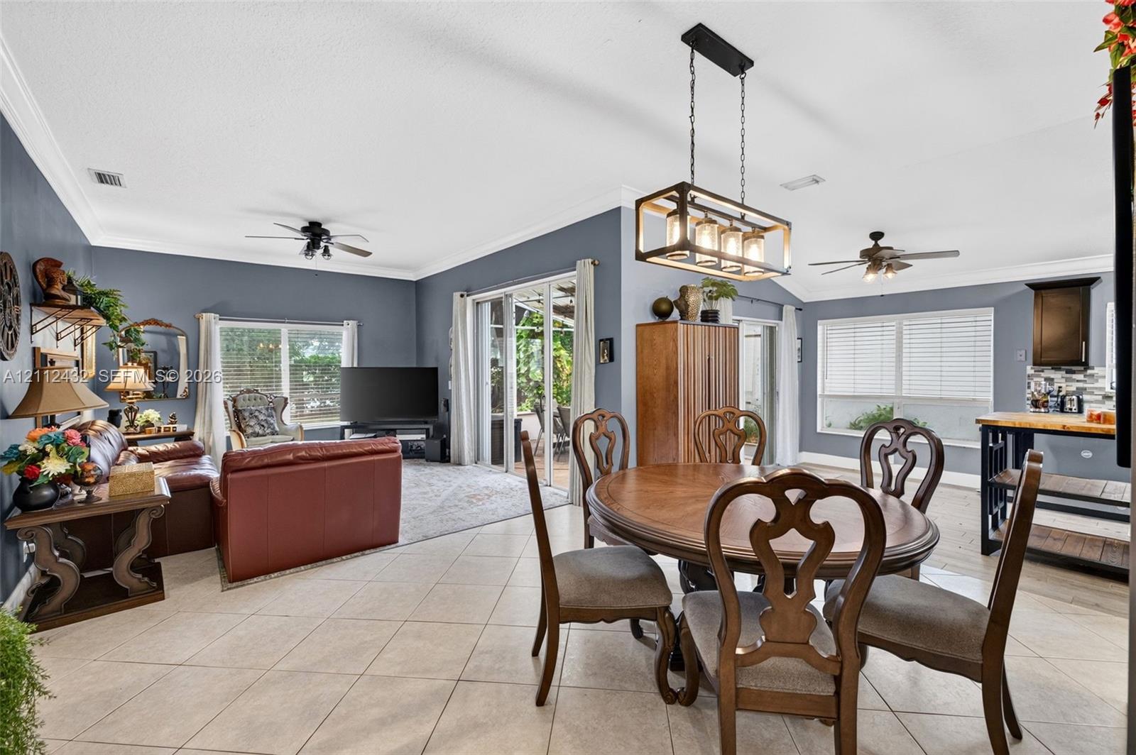 Open-concept main area featuring large-format ceramic tile flooring, crown molding, multi-light rectangular pendant fixture, dual ceiling fans, and multi-panel sliding glass doors