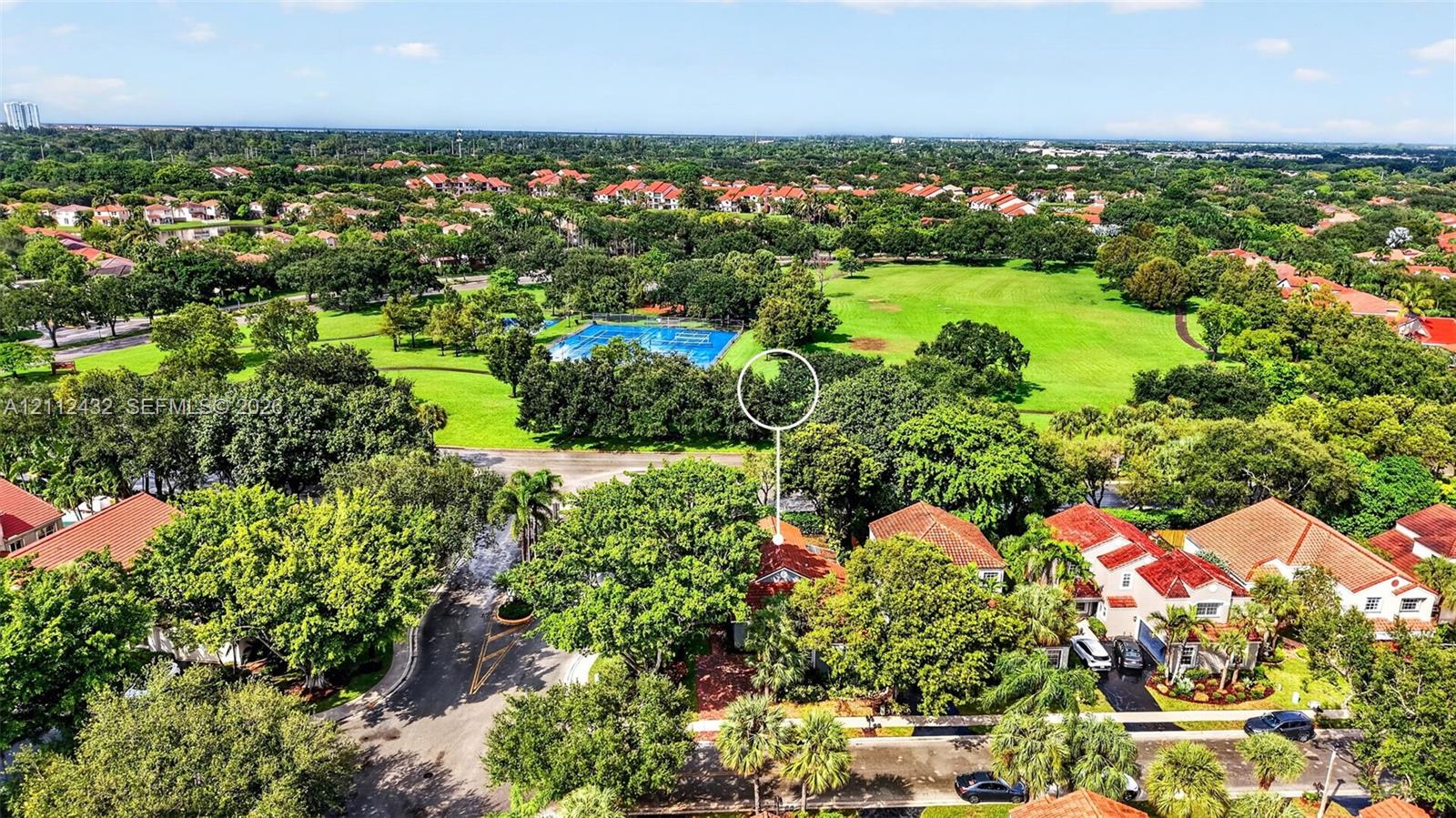 Aerial perspective featuring a residential lot with mature trees, a red-tiled roof, and proximity to expansive green space and a community recreational area