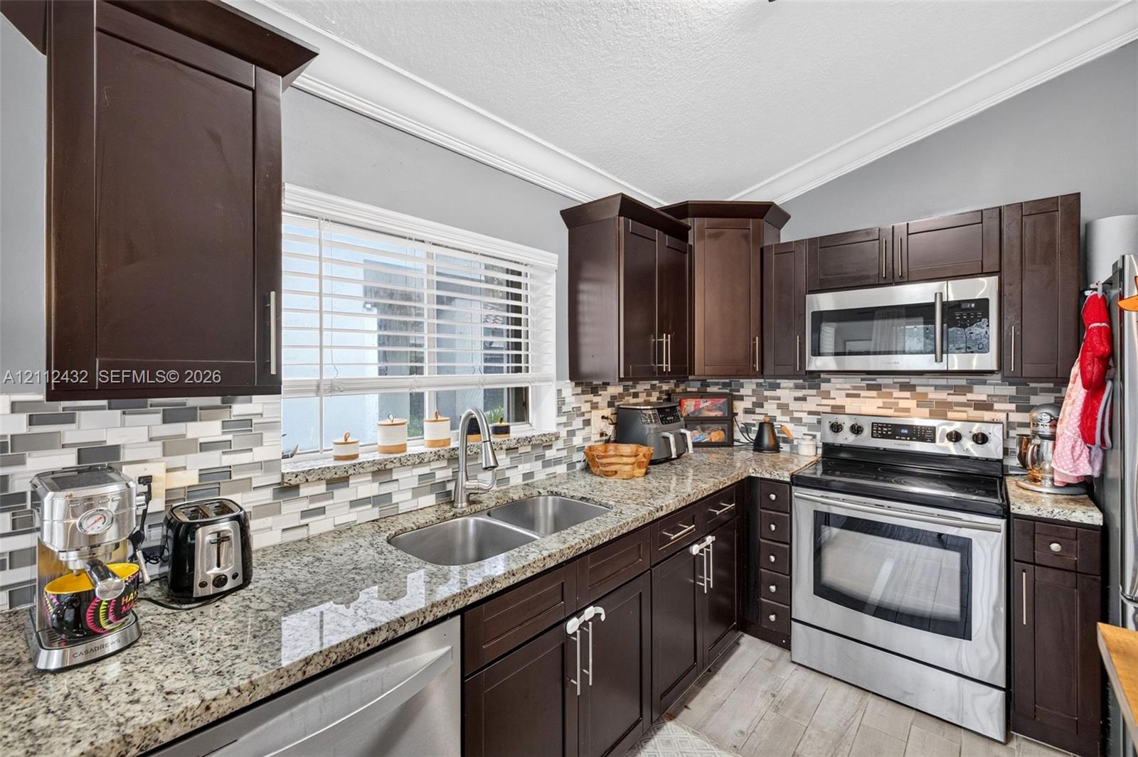 Kitchen featuring dark wood cabinetry, granite counters, stainless appliances, a multi-tone tile backsplash, and wood-look tile flooring