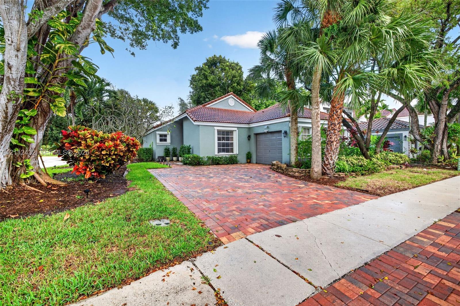 Single-story home featuring a 2015 tile roof, 2025 paver driveway, attached garage, and beautiful established landscaping with mature palm trees