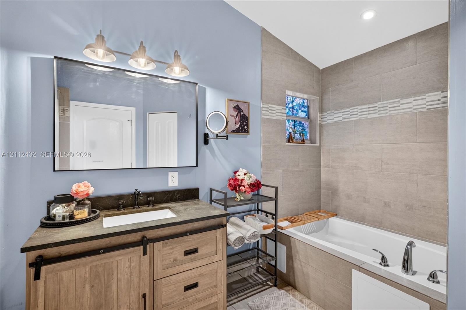 Wood-look vanity with a dark counter and rectangular drop-in sink, framed mirror with three-bulb vanity lighting, and a bathtub with tile surround and separate shower