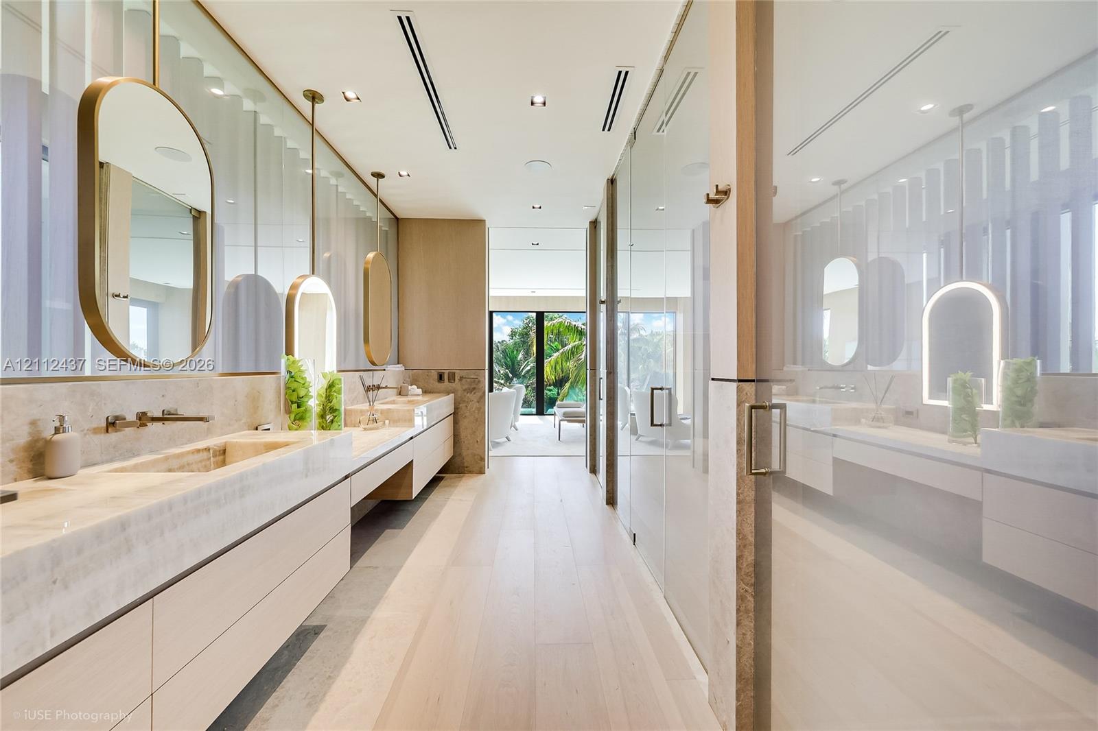 Dual floating vanities with integrated stone sinks and wall-mounted faucets, suspended oval mirrors, recessed ceiling lighting, wood-finish flooring, and floor-to-ceiling glass paneling