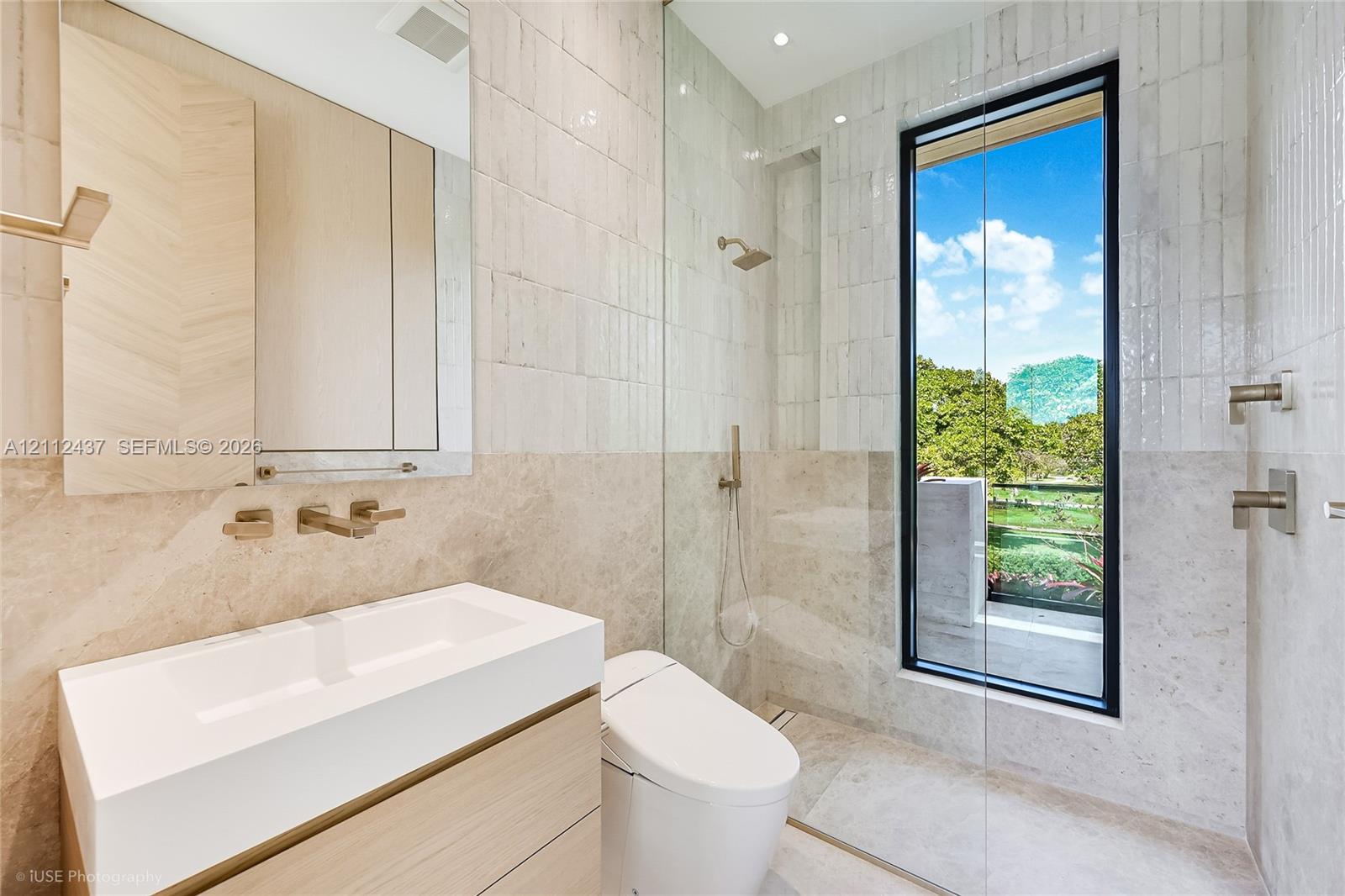 Modern bathroom featuring a wood-finish floating vanity, wall-mounted metallic fixtures, a frameless glass shower enclosure, large-format stone tiling, and a floor-to-ceiling window