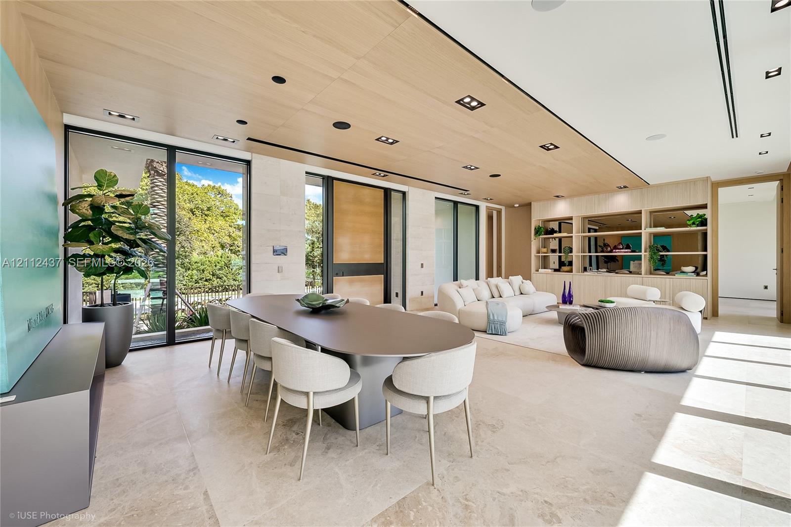 Modern open-concept interior featuring light wood-finish cabinetry, floor-to-ceiling glass sliding walls, stone-finished kitchen island, recessed lighting, and large-format tile flooring Modern open-concept interior featuring light wood-finish cabinetry, floor-to-ceiling glass sliding walls, stone-finished kitchen island, recessed lighting, and large-format tile flooring