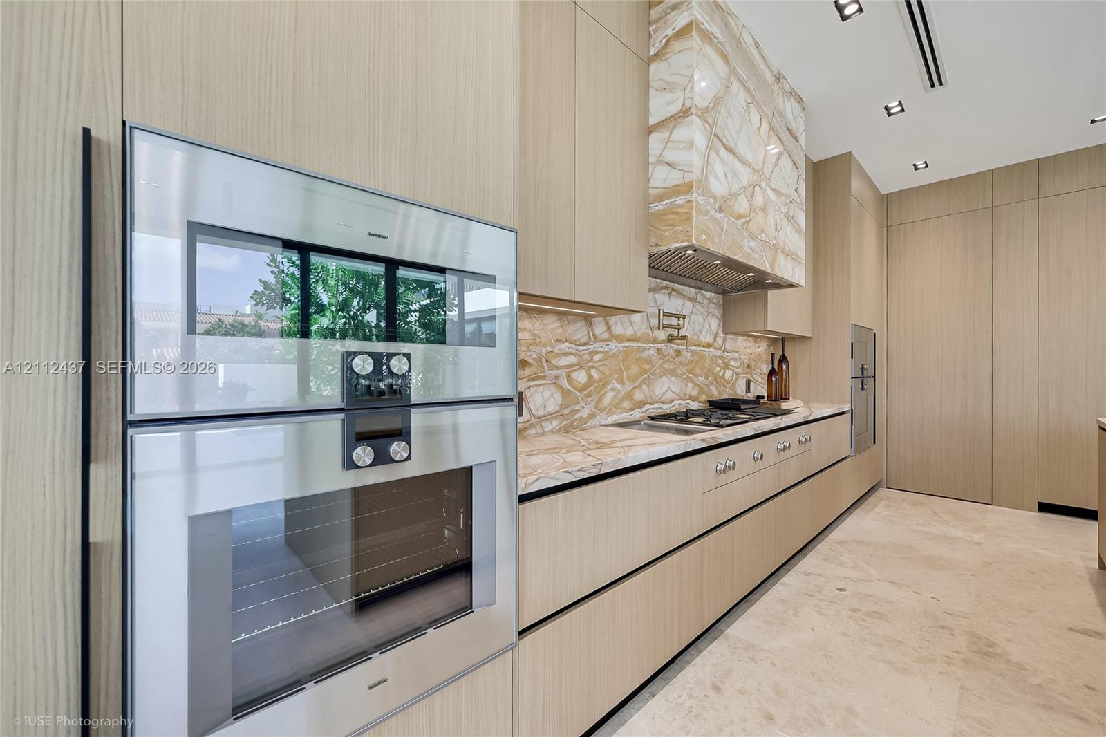 Modern kitchen featuring flat-panel wood-finish cabinetry, floor-to-ceiling stone-slab backsplash and hood surround, integrated gas cooktop with pot filler, expansive stone-topped island, and oversized black-framed glass sliding doors