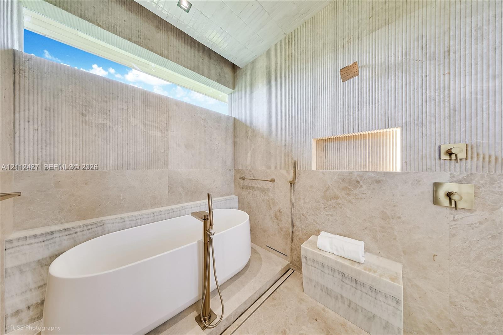 Freestanding white soaking tub with a floor-mounted faucet, floor-to-ceiling stone wall tiling, integrated recessed niche with accent lighting, built-in stone bench, and horizontal clerestory window