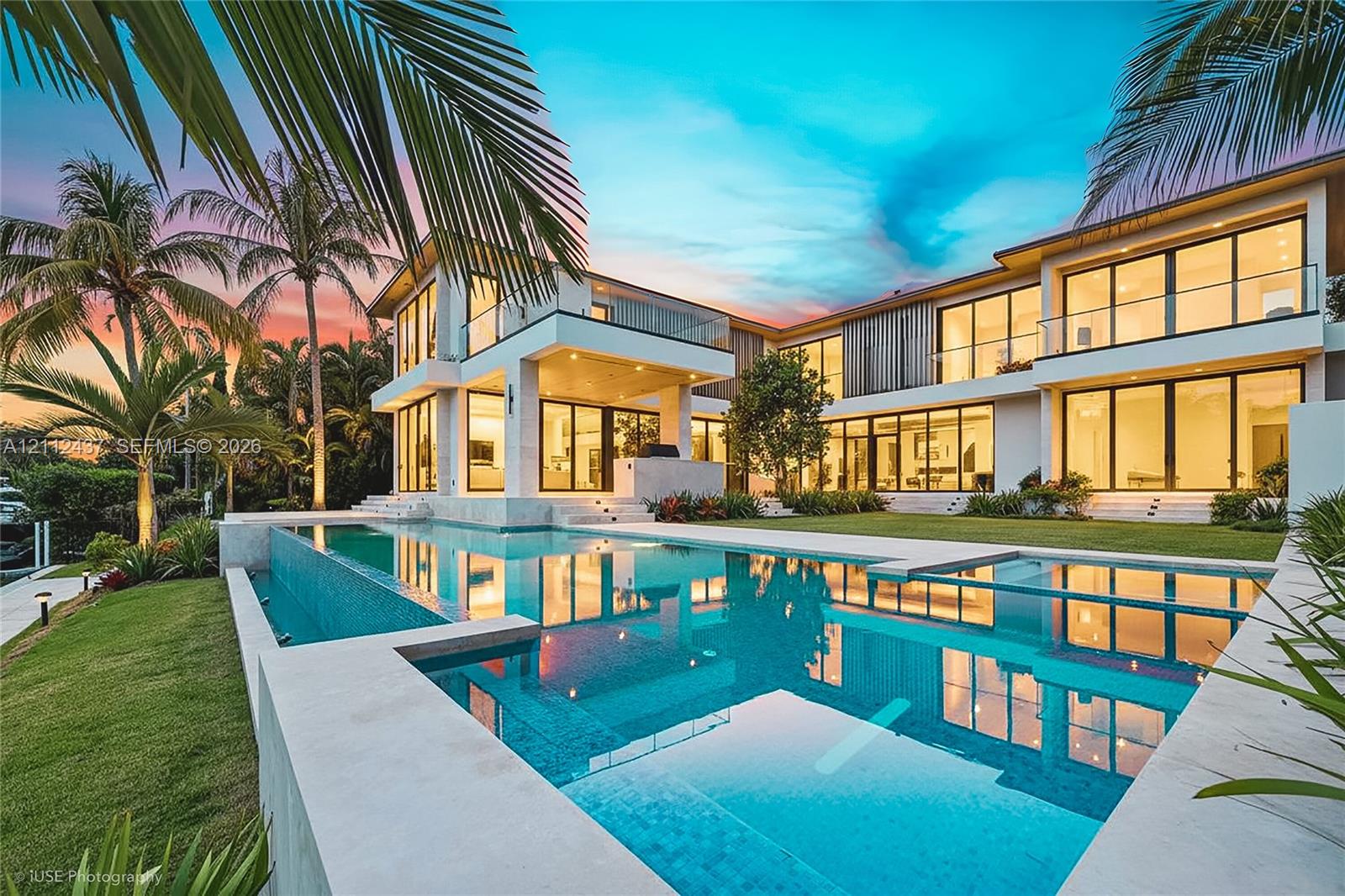 Two-story contemporary residence featuring expansive floor-to-ceiling glass walls, an infinity-edge pool, multiple covered balconies with glass railings, and stone pool decking