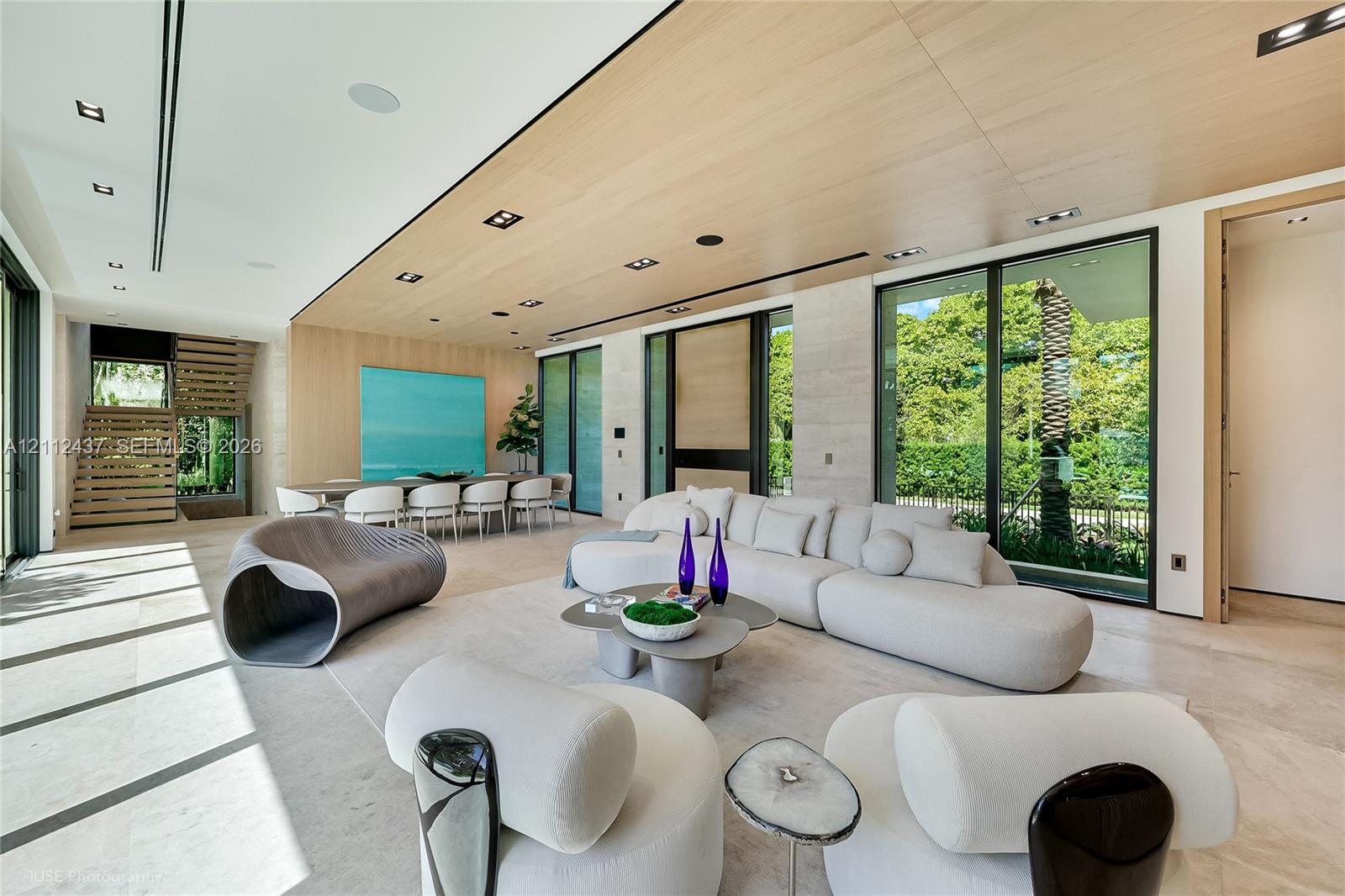 Open-concept living space featuring wood-clad ceilings, floor-to-ceiling glass sliding doors, stone-finish flooring, and built-in light-tone wood shelving Open-concept living space featuring wood-clad ceilings, floor-to-ceiling glass sliding doors, stone-finish flooring, and built-in light-tone wood shelving
