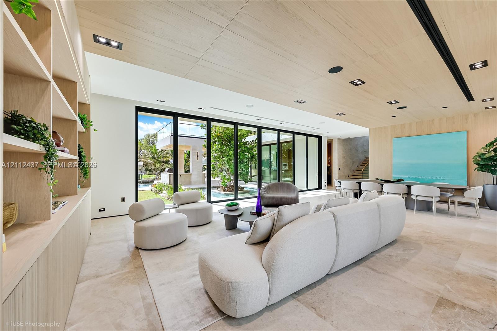 Open-concept interior featuring natural stone flooring, expansive floor-to-ceiling glass sliding doors, wood-paneled ceiling with recessed lighting, and custom built-in wall shelving Open-concept interior featuring natural stone flooring, expansive floor-to-ceiling glass sliding doors, wood-paneled ceiling with recessed lighting, and custom built-in wall shelving