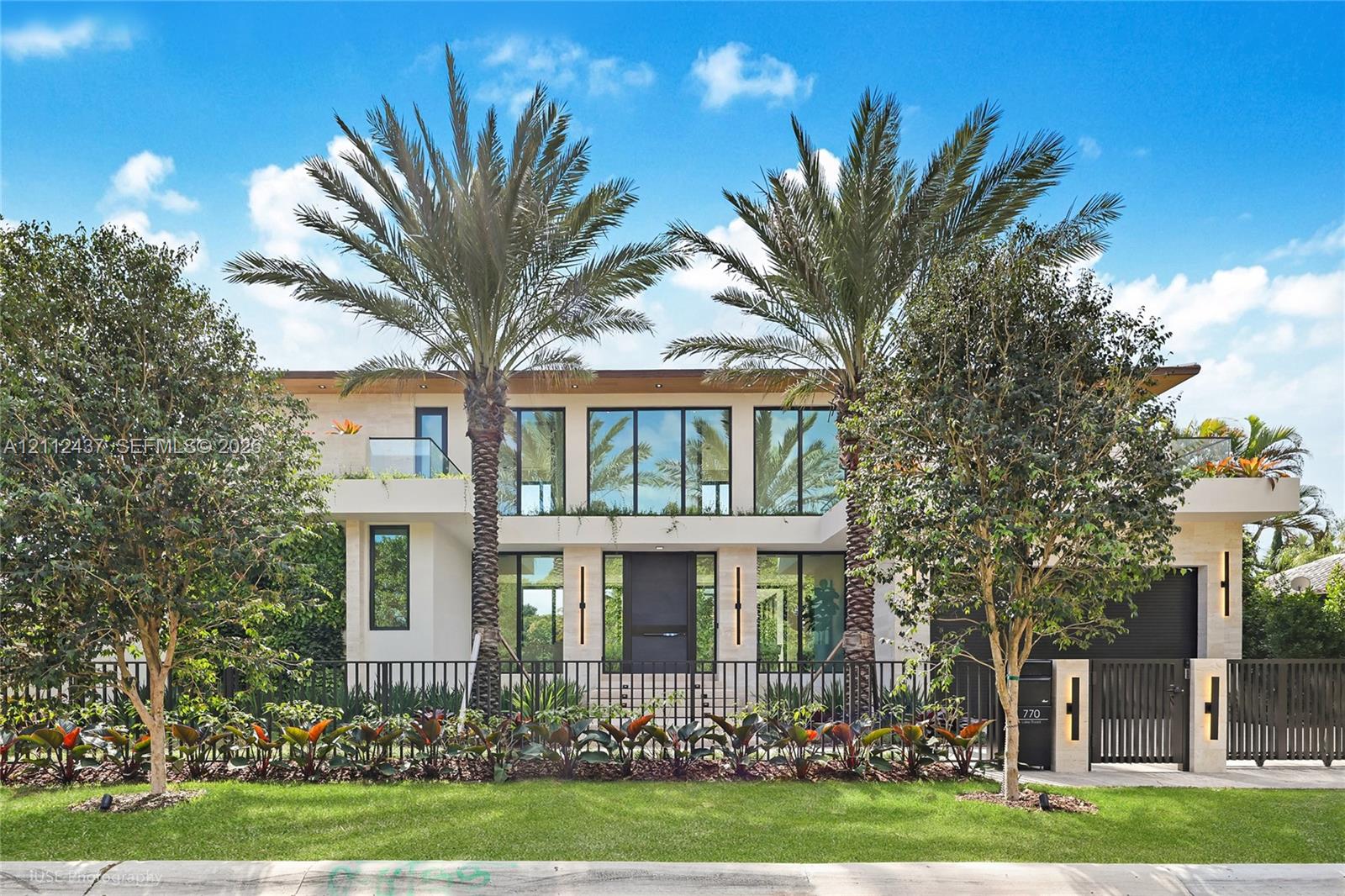Contemporary two-story exterior featuring smooth stucco facade, floor-to-ceiling glass windows, integrated wall-mounted lighting, manicured landscaping, and decorative metal fencing