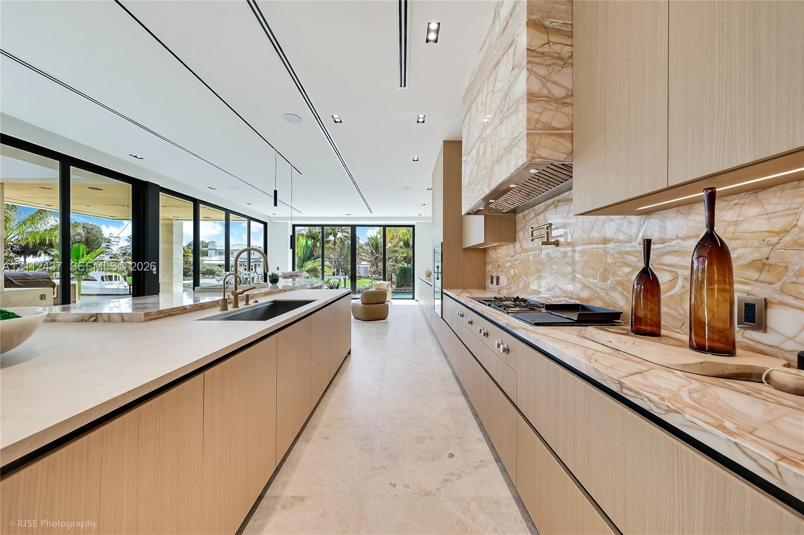 Custom wood-finish cabinetry, expansive stone-topped kitchen island with waterfall edge, integrated stainless steel appliances, and floor-to-ceiling glass sliding doors Custom wood-finish cabinetry, expansive stone-topped kitchen island with waterfall edge, integrated stainless steel appliances, and floor-to-ceiling glass sliding doors