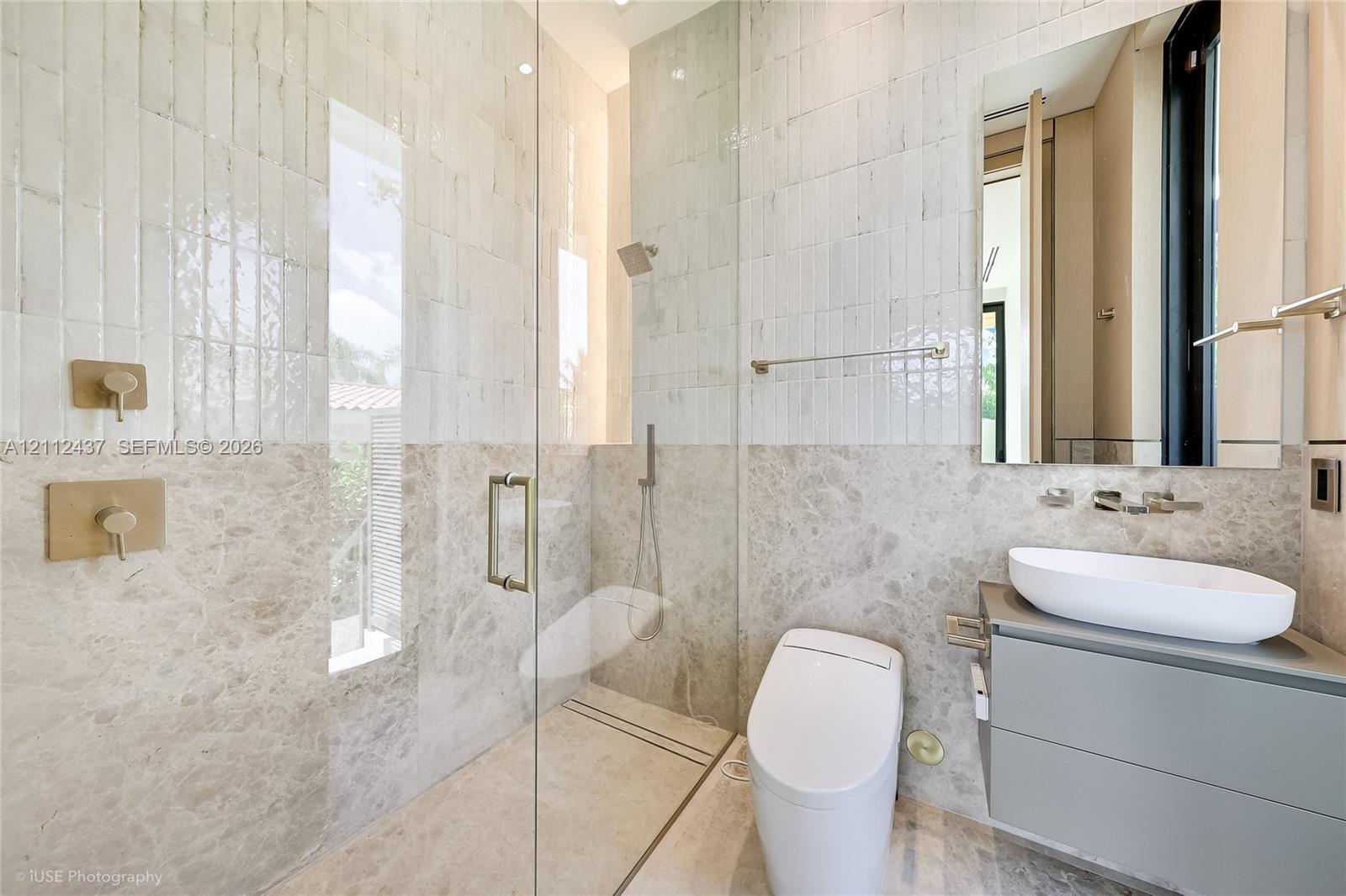 Full-height tiled walk-in shower with a glass enclosure, wall-mounted brass fixtures, stone-finish surround, floating vanity with a vessel sink, and one-piece low-profile toilet