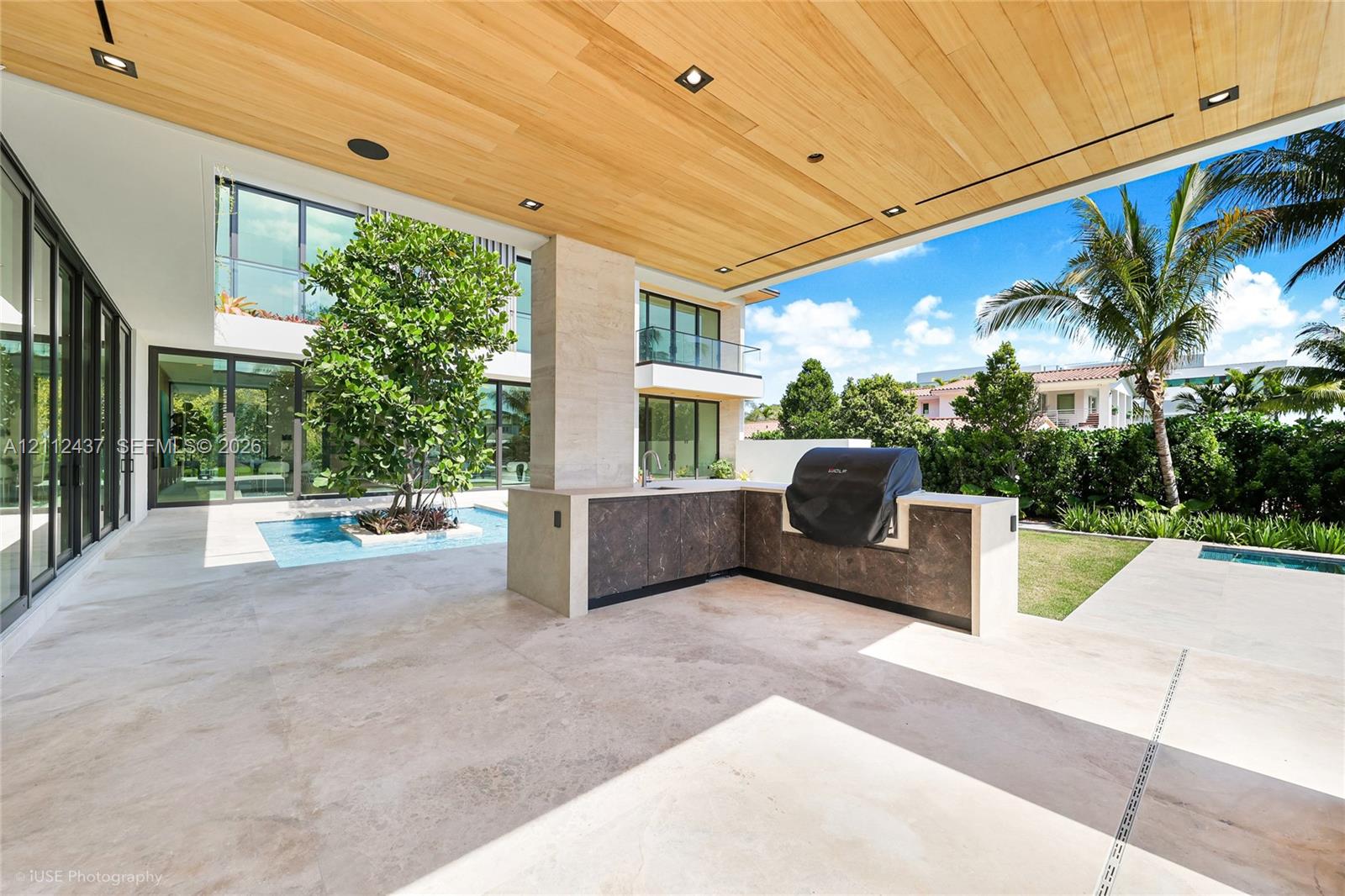 Covered outdoor living space featuring a wood-plank ceiling, natural stone tile flooring, integrated outdoor kitchen with stone countertop, and a centered water feature