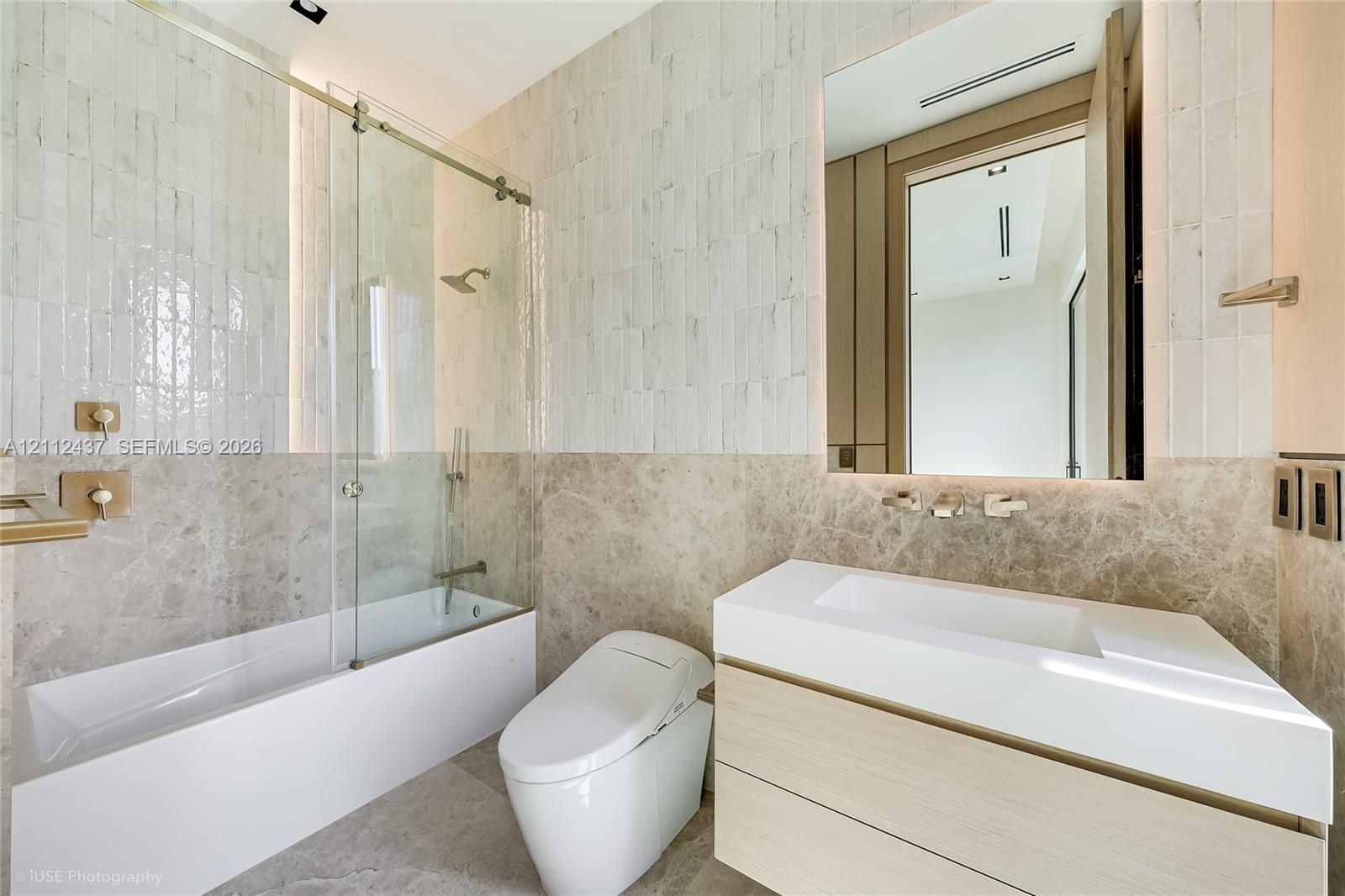 Bathroom featuring a floating vanity with a white countertop, wall-mounted faucet, glass-enclosed soaking tub with stone tile surround, and textured wall tile