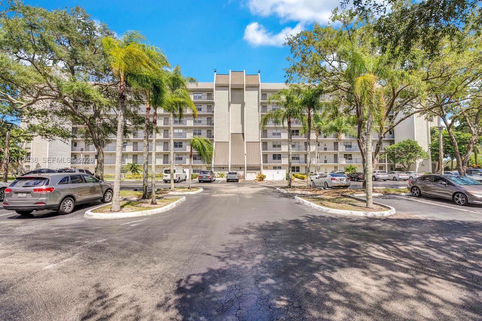 Multi-story residential building featuring a textured facade, structural concrete elements, private balconies, on-site parking, and mature palm trees