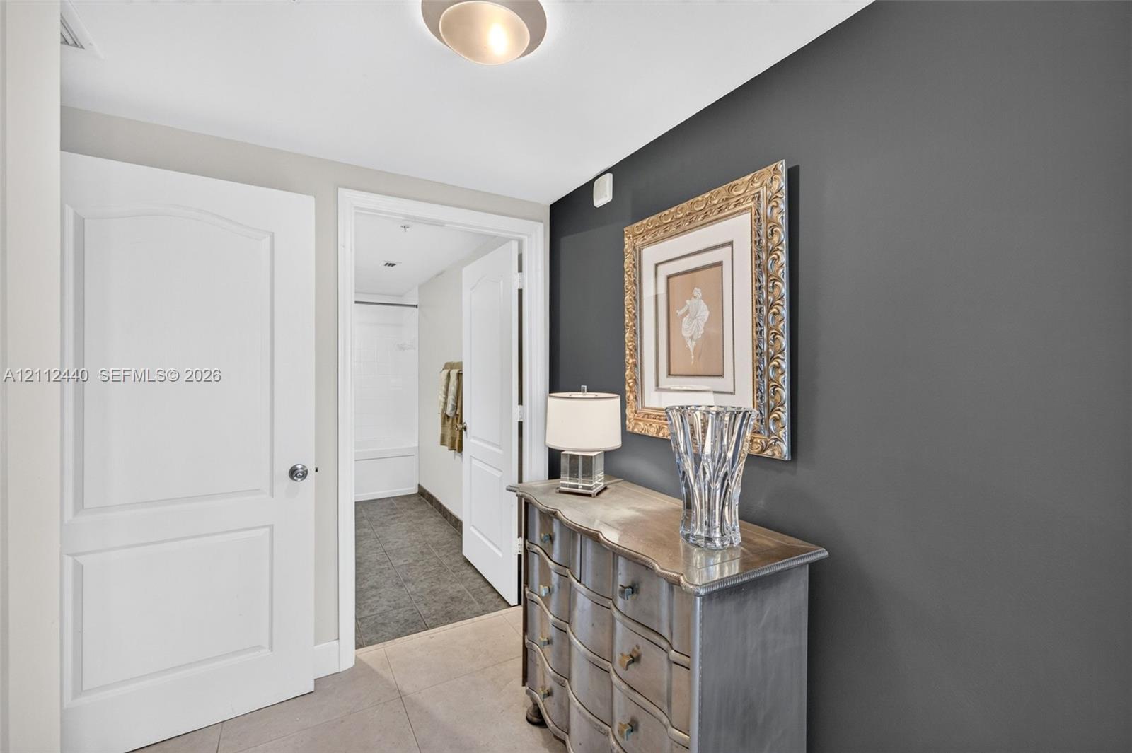 Entrance to primary bedroom and ensuite bathroom, featuring a charcoal accent wall, neutral-toned porcelain flooring, flush-mount ceiling lighting