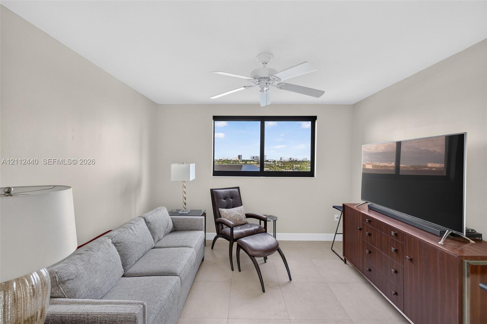 Neutral-toned guest bedroom featuring large-format porcelain flooring, a central ceiling fan with integrated lighting, and impact window with motorized roller shade