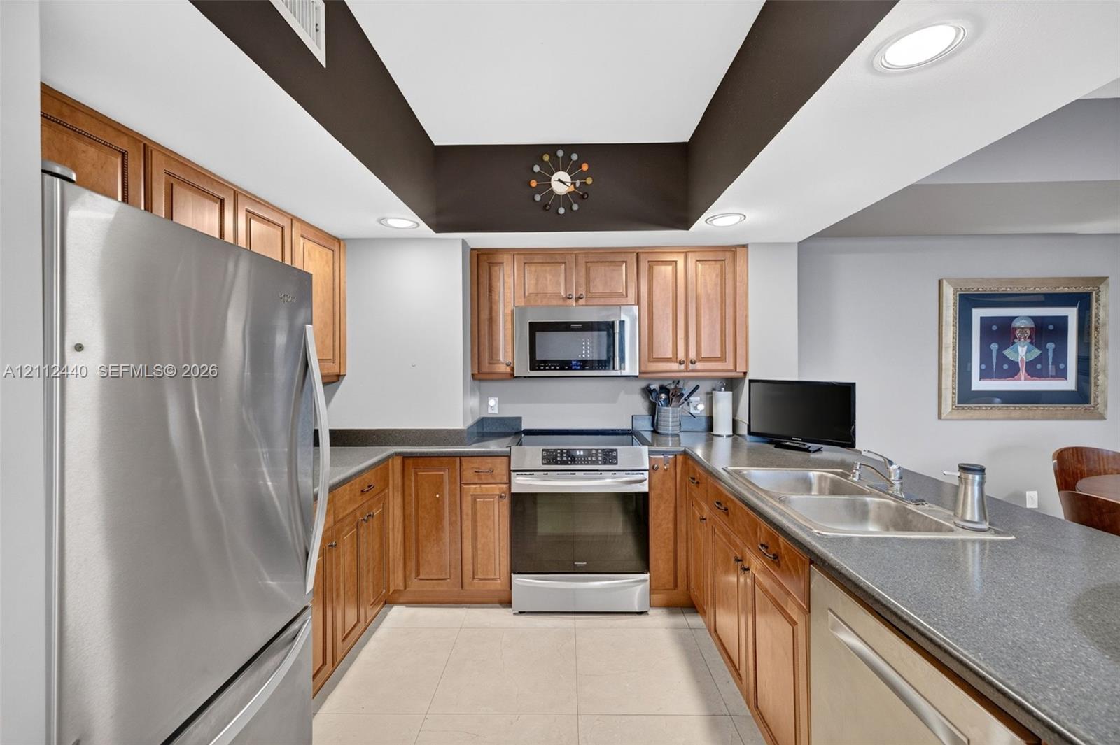 Kitchen featuring recessed tray ceiling, wood cabinetry, stainless appliances, dual-basin stainless steel sink, and dark countertops