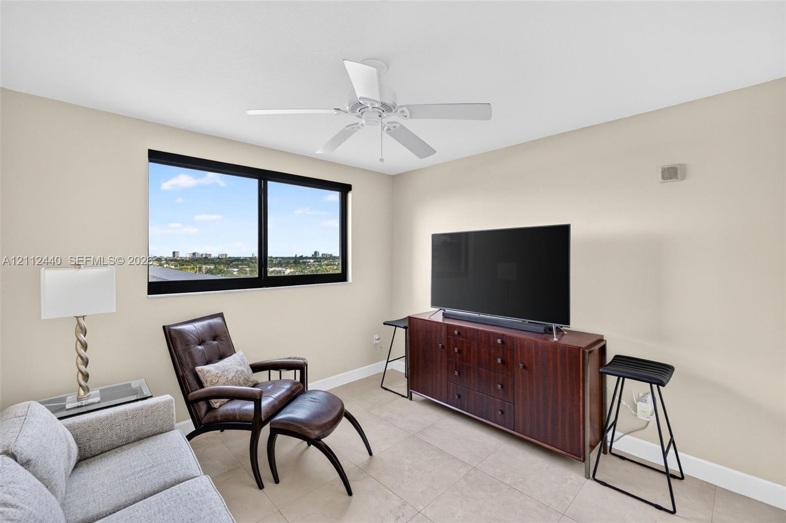 Neutral-toned room featuring a large window with black framing, tile flooring, and a white ceiling fan with integrated lighting