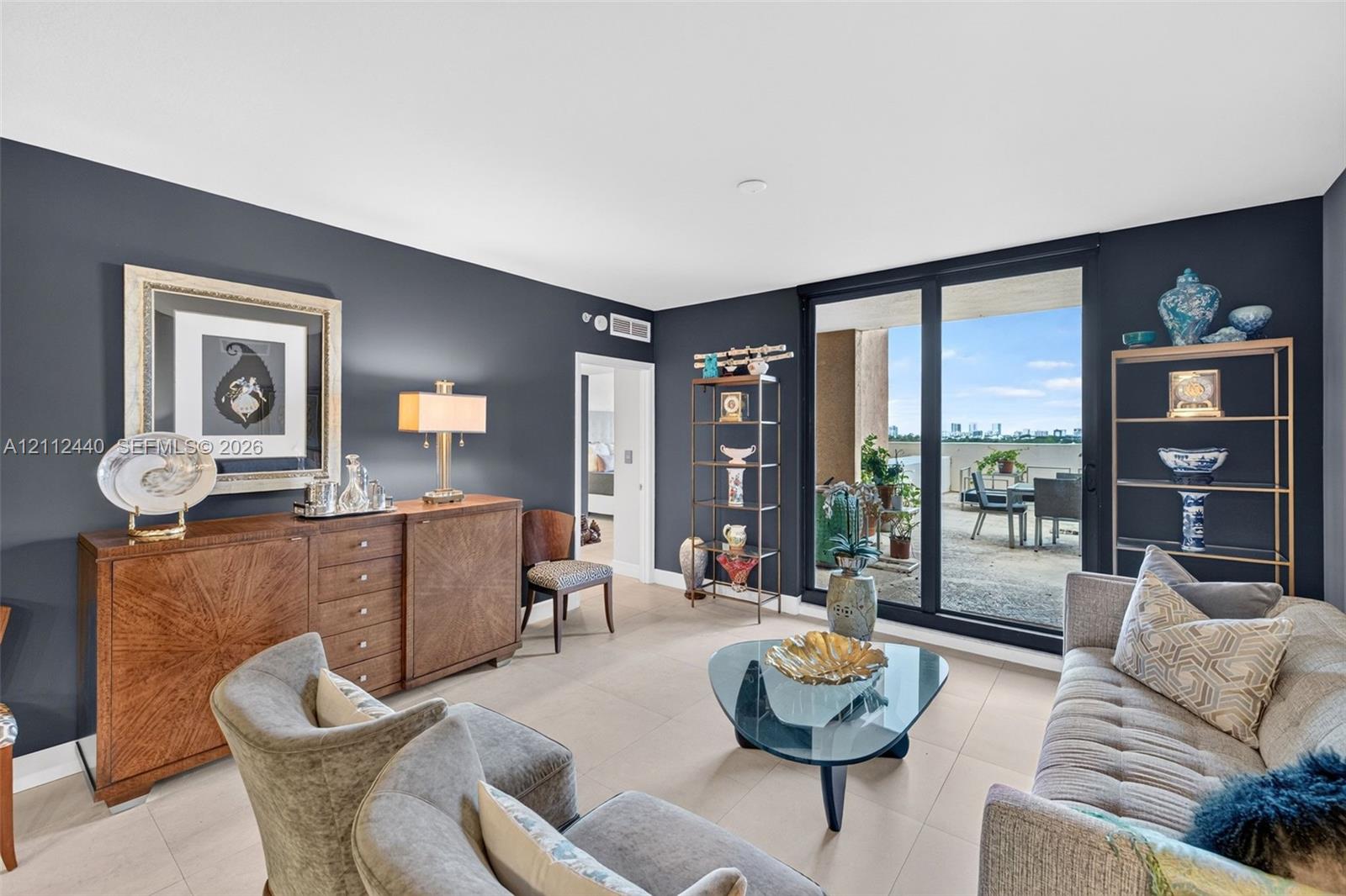 Living area with light-toned porcelain flooring, deep-toned accent walls, and floor-to-ceiling sliding glass doors opening to a huge concrete balcony w/ condo views by the beach