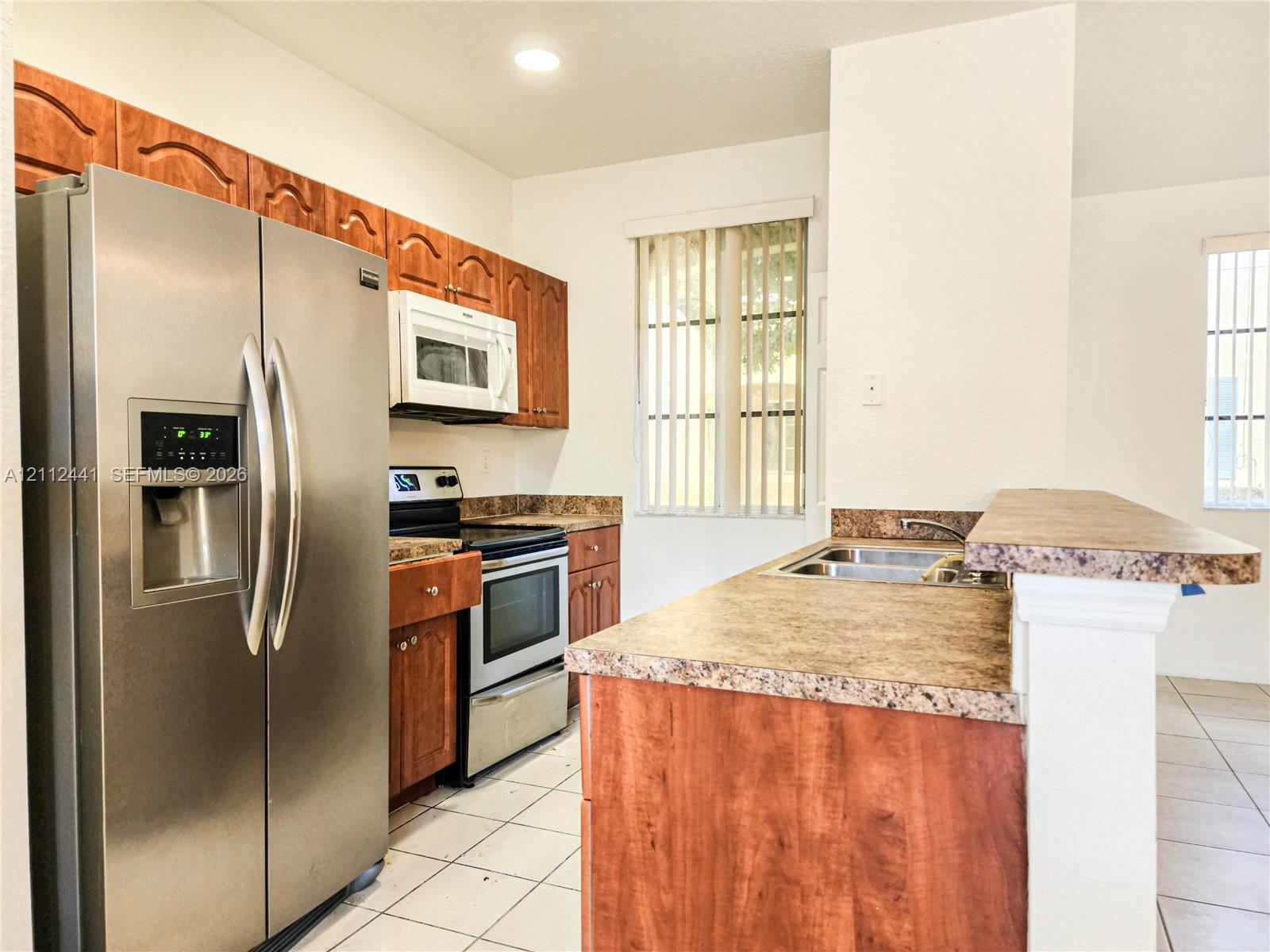 Kitchen featuring stainless steel appliances, wood cabinetry, patterned laminate countertops, a dual-basin sink, and tile flooring Kitchen featuring stainless steel appliances, wood cabinetry, patterned laminate countertops, a dual-basin sink, and tile flooring