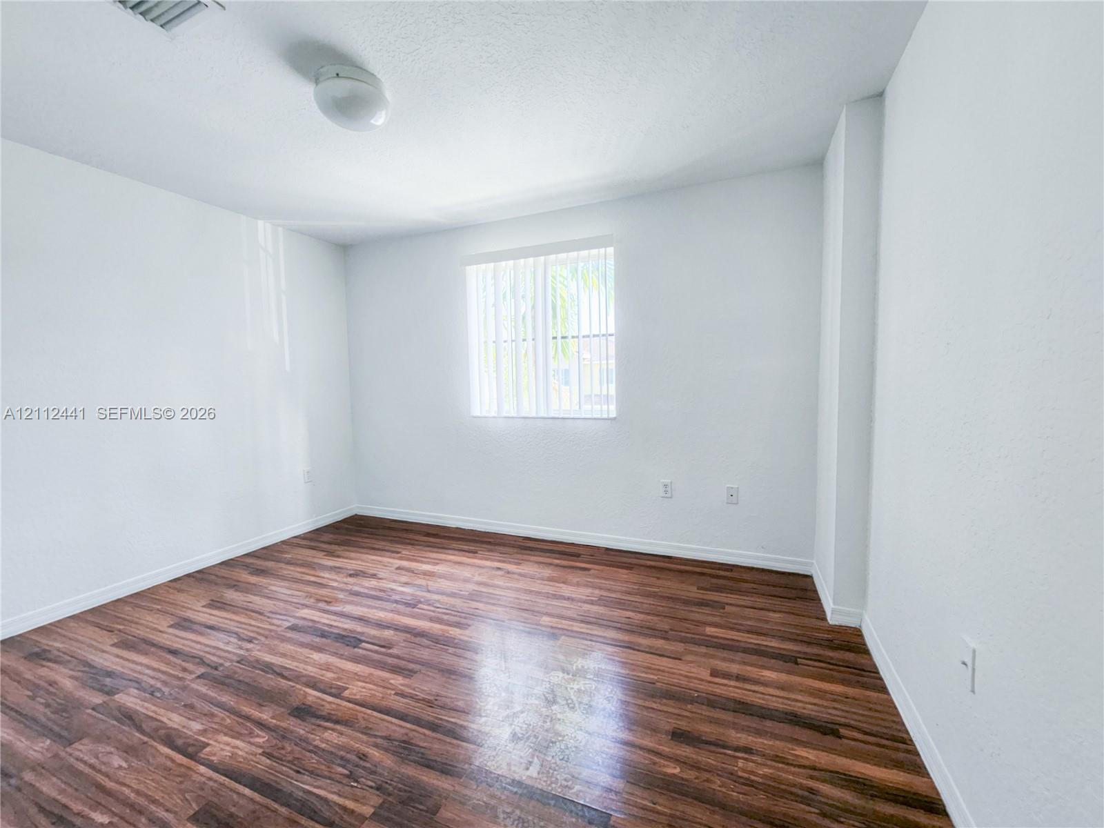 Room featuring wood-finish flooring, white walls, and a single window with vertical blinds Room featuring wood-finish flooring, white walls, and a single window with vertical blinds