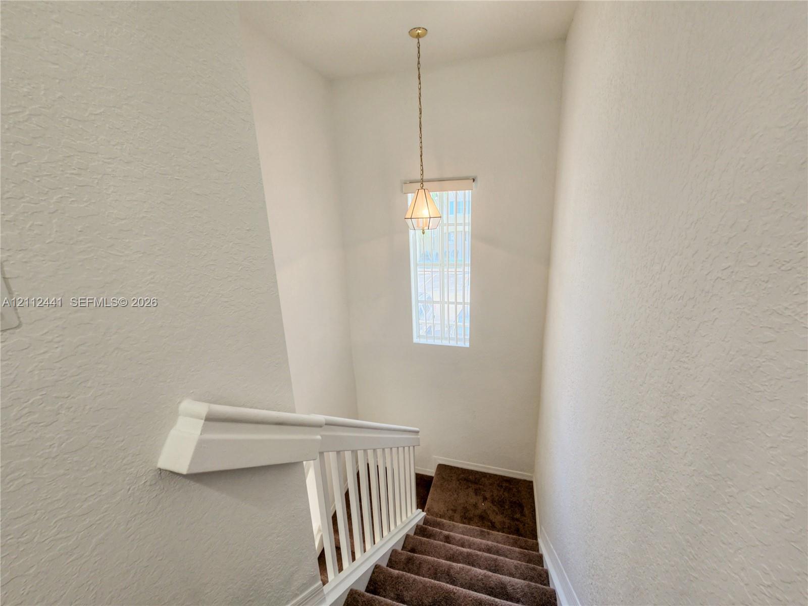 Stairwell featuring carpeted steps, white baluster railing, and a geometric pendant light fixture Stairwell featuring carpeted steps, white baluster railing, and a geometric pendant light fixture