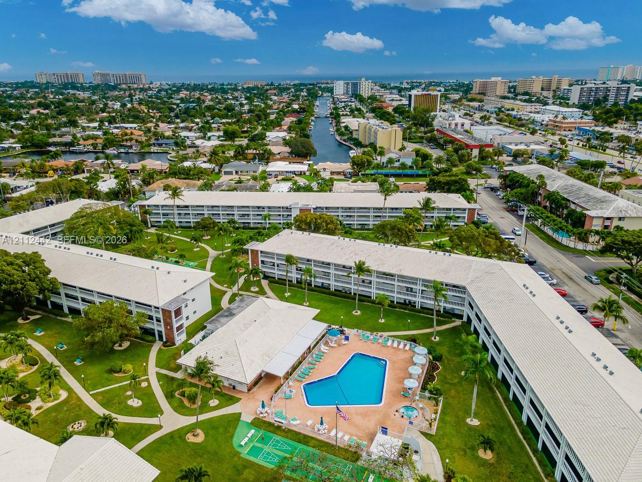 Multi-story residential complex featuring a central outdoor swimming pool, spa, manicured landscaping, paved walkways, and a dedicated shuffleboard court