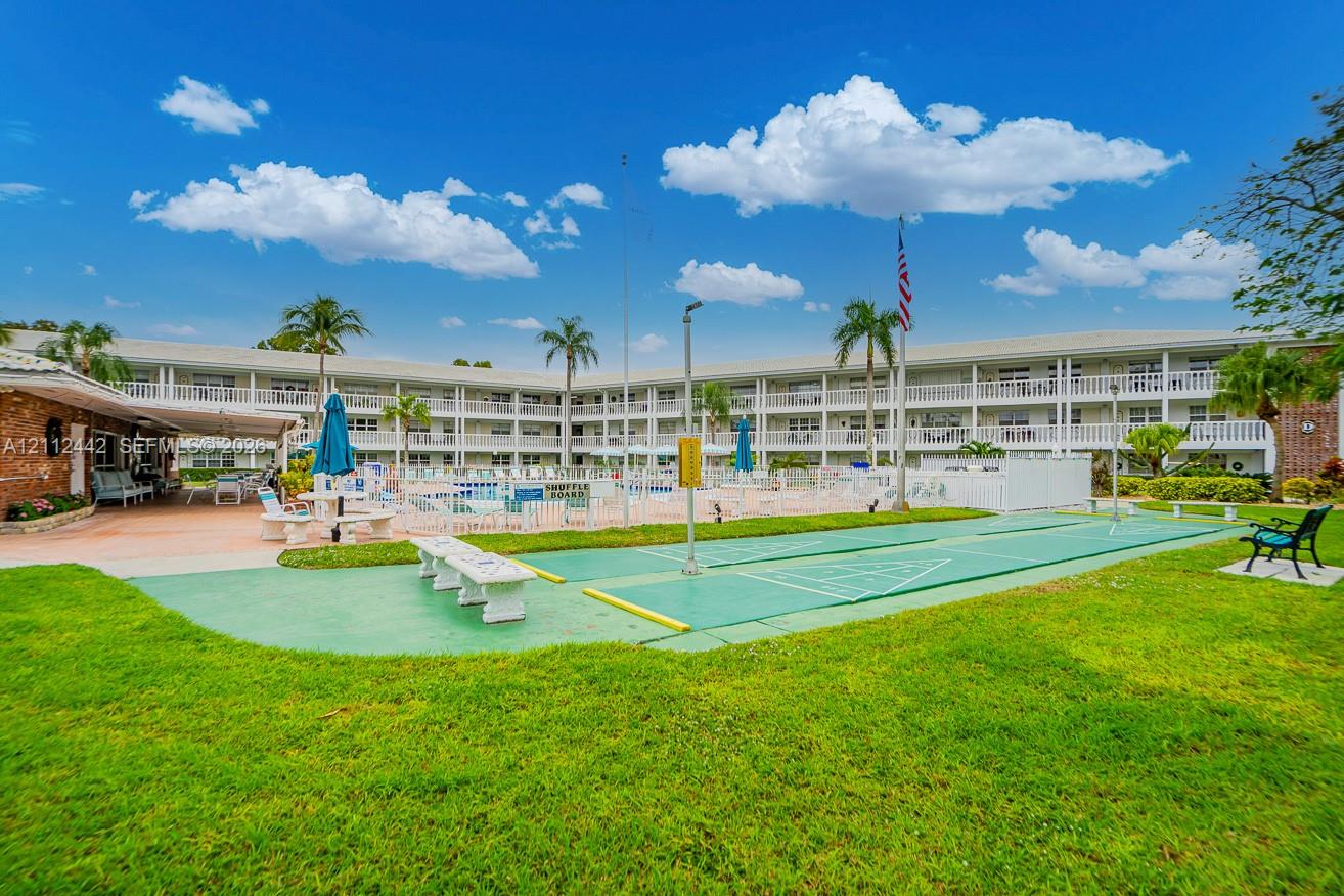 Outdoor recreation area featuring a shuffleboard court, a swimming pool with enclosure, a landscaped lawn, and palm trees