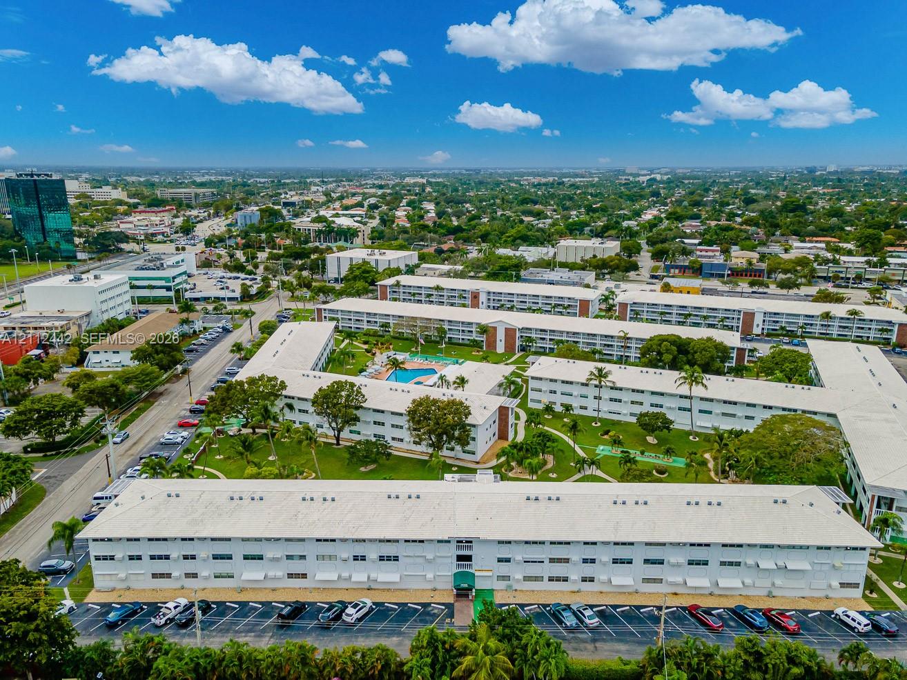 Aerial perspective of a multi-building residential complex featuring white exterior facades, an in-ground swimming pool, manicured grounds, and dedicated surface parking