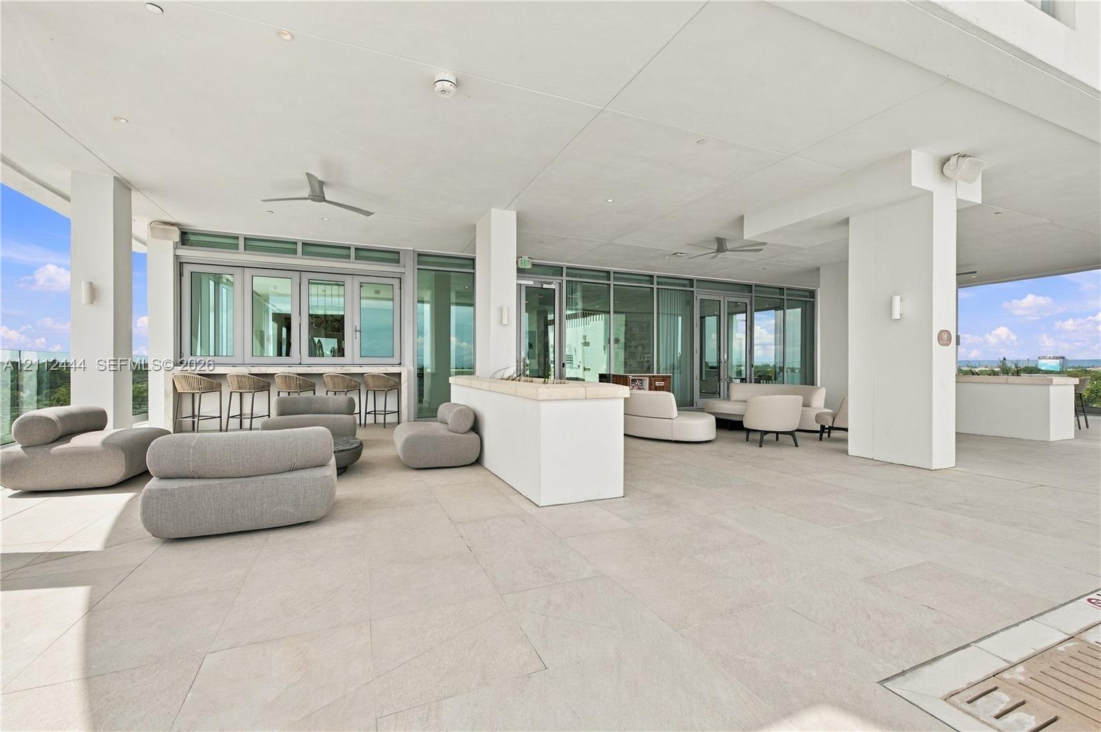Covered terrace featuring large-format stone tile flooring, built-in stone bar counter, floor-to-ceiling glass paneling, and ceiling-mounted fans